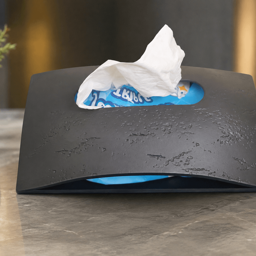 A black textured tissue box cover or tissue dispenser sits on a gray stone countertop, with a white tissue pulled out from a blue tissue box visible inside. A small potted plant is blurred on the left, and the background is softly lit in warm neutral tones.